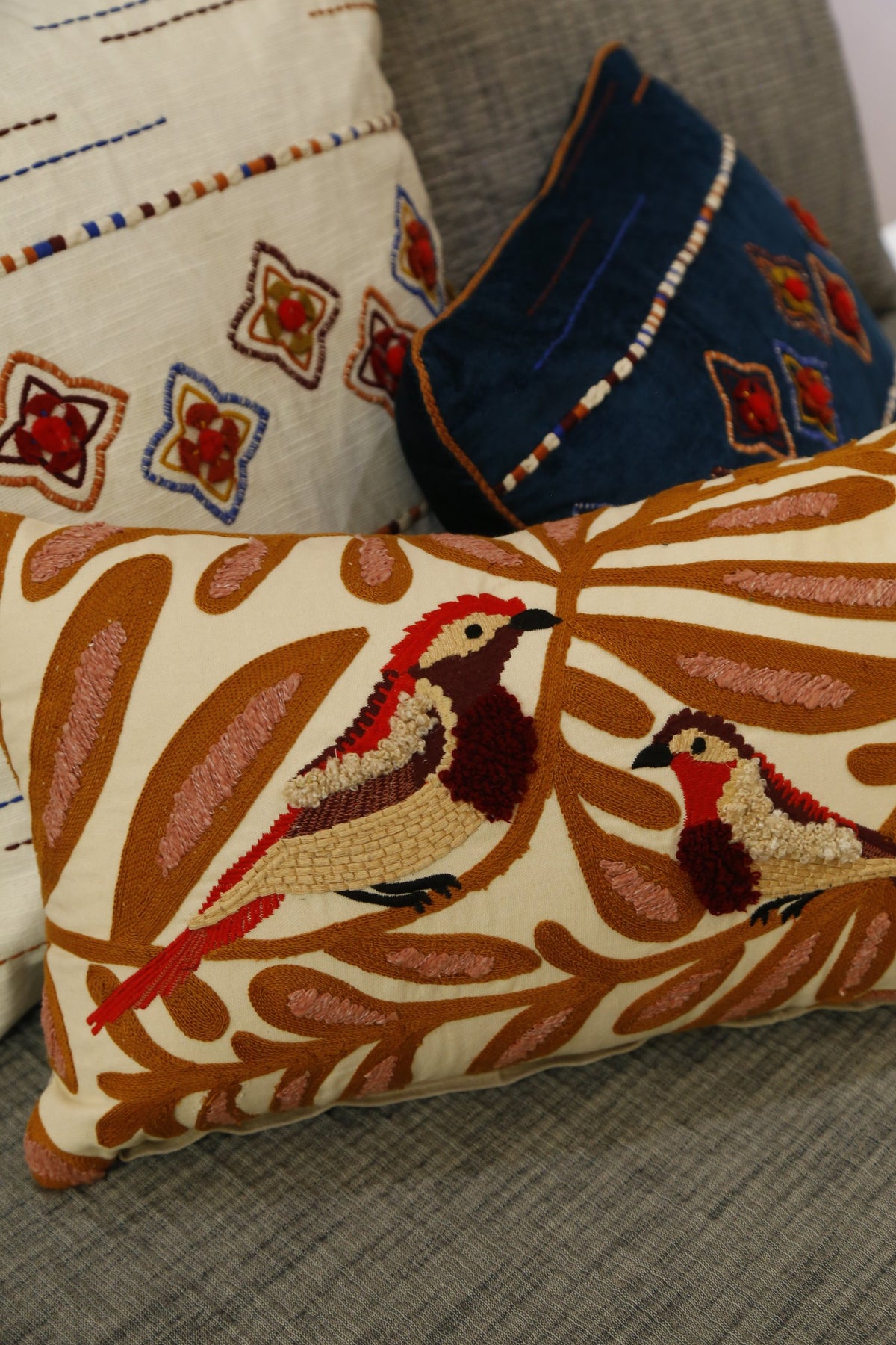 Multi bird and vine embroidered cushion cover, angled view on a grey bed